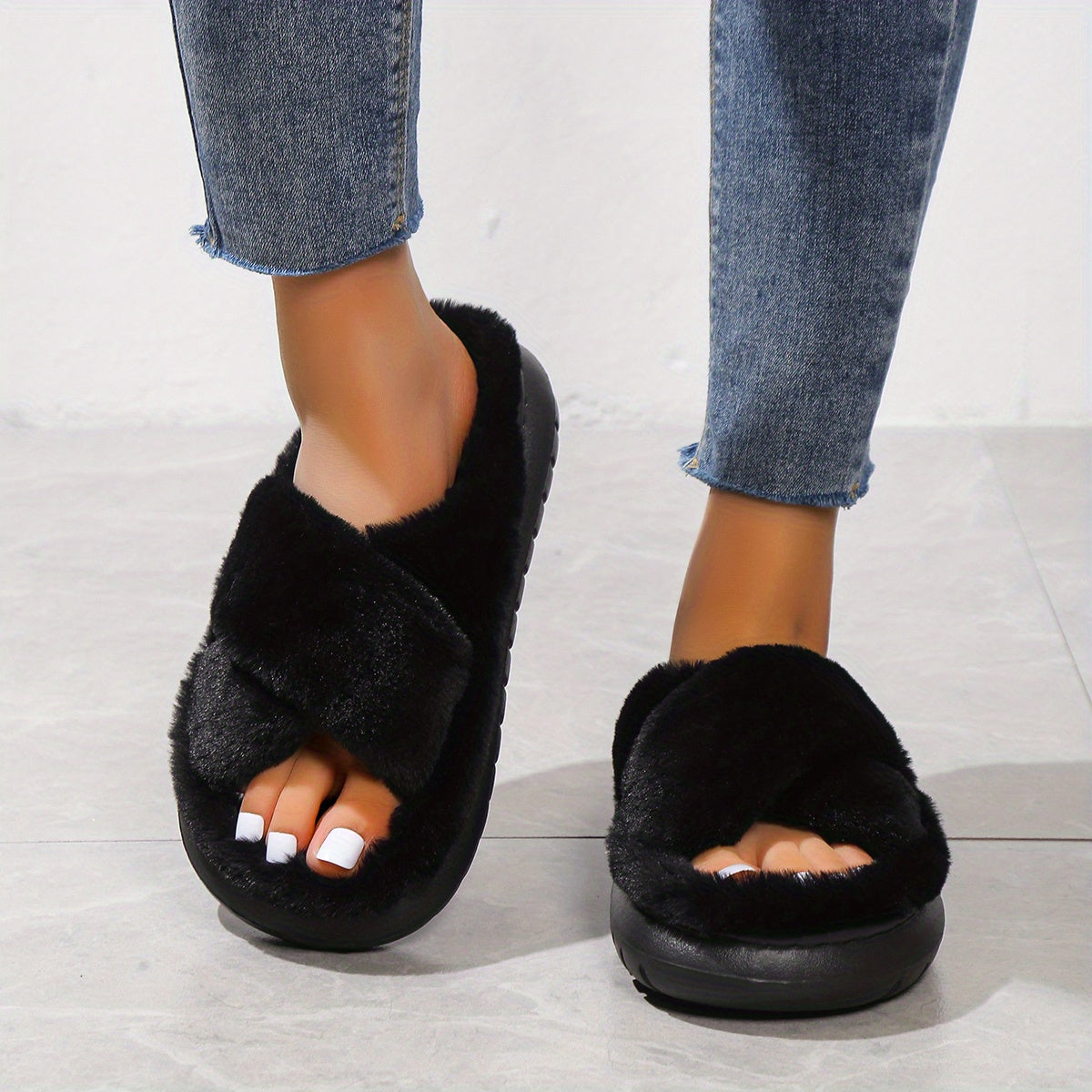 Black fuzzy slippers worn with blue jeans on a light wooden floor.