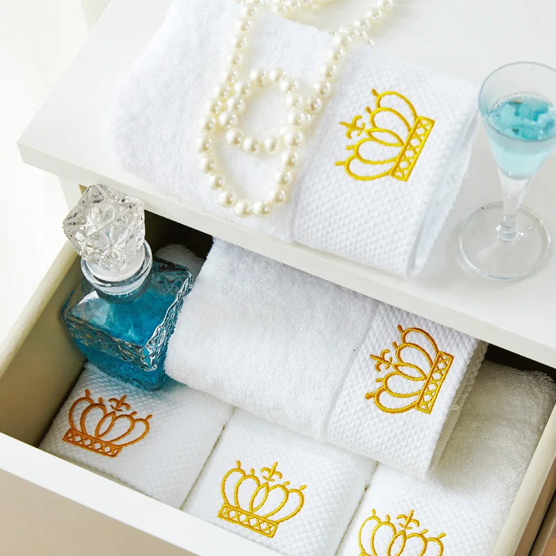 White towels with gold crowns, a bottle of blue perfume, and a glass with blue liquid on a white surface.