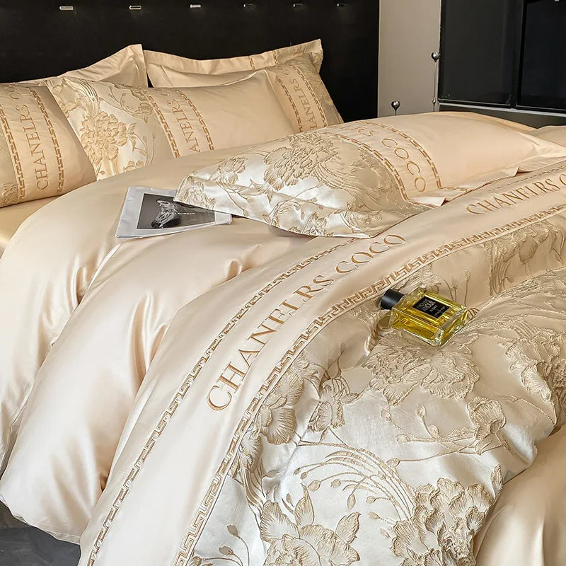 Luxurious beige bedding set with floral patterns and 'Chanel' branding, featuring a bottle of perfume on top.