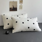 Two white pillows with black polka dots on a gray surface, with a light gray wall and framed picture in the background.