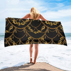 Person holding a black beach towel with gold chain pattern on a beach.